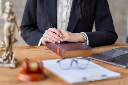 Person in suit sitting at desk with a book, pen, laptop, glasses, and legal statue.