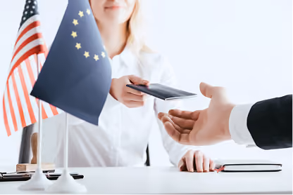 Person handing a passport to another across a desk with American and European Union flags.