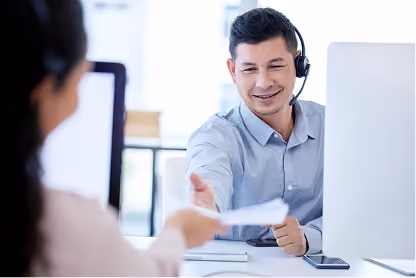 Man in headset at desk handing a document to a woman across the table in a bright office.