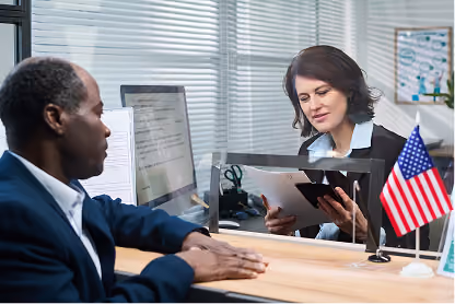 Man and woman separated by clear divider, woman reviewing documents at a desk with US flag.
