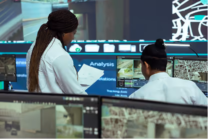 Two professionals review data and maps on large computer screens in a control or operations room.