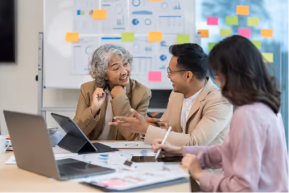 Three professionals discussing work with charts and sticky notes on the board in a meeting room.