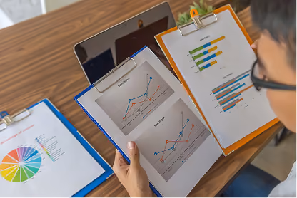 Person reviewing clipboards with colorful charts and graphs next to a laptop on a wooden table.
