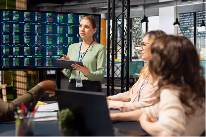 Woman in green blouse presenting stock market data on screen to colleagues in an office meeting.