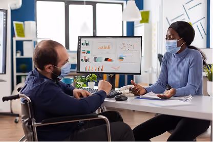 Two masked colleagues discuss data charts displayed on a computer monitor in a modern office.