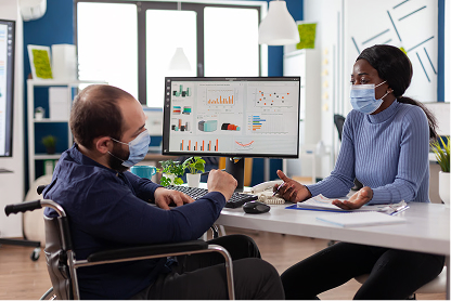 Two masked colleagues discuss data charts displayed on a computer monitor in a modern office.