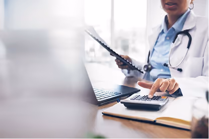 Doctor using a calculator and holding a clipboard at a desk with a laptop and notebook.
