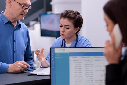 A healthcare worker discusses information with a man at a reception desk while another person uses a phone.