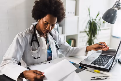 Female doctor in white coat reviews papers while sitting at desk with laptop in bright office.