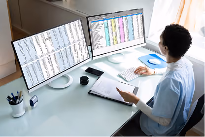 Person analyzing data on dual computer monitors with spreadsheets while holding documents at a desk.
