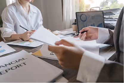 Two people reviewing financial documents and reports at a desk with a laptop and coffee cup.