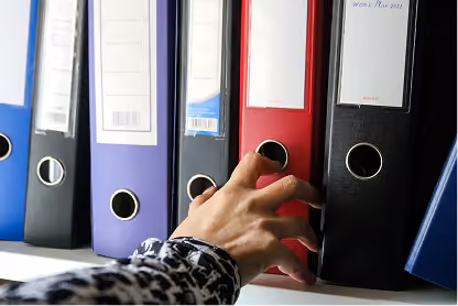 Hand reaching for a black binder on a shelf among several colored binders.