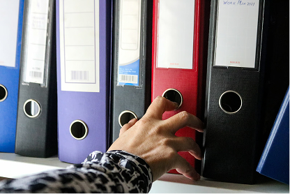 Hand reaching for a black binder on a shelf among several colored binders.