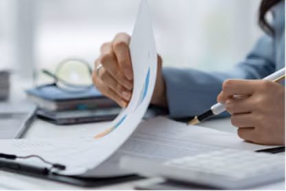 Person reviewing documents with a pen in hand on a desk with glasses and a laptop nearby.