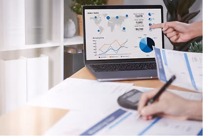 Person pointing at laptop screen showing graphs and charts while another writes on paper with calculator nearby.