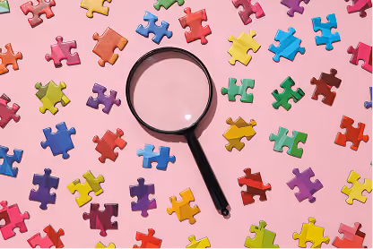 Magnifying glass on pink surface surrounded by scattered colorful jigsaw puzzle pieces.