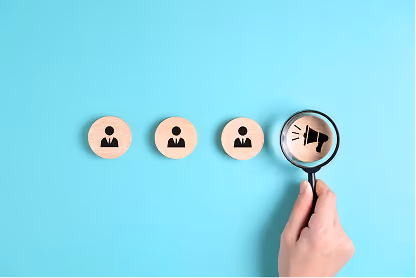 Hand holding magnifying glass over wooden icon of megaphone next to three user icons on blue background.