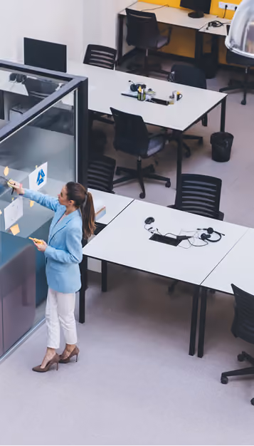 Businesswoman in blue blazer writing on glass wall in a modern office with empty desks and chairs.