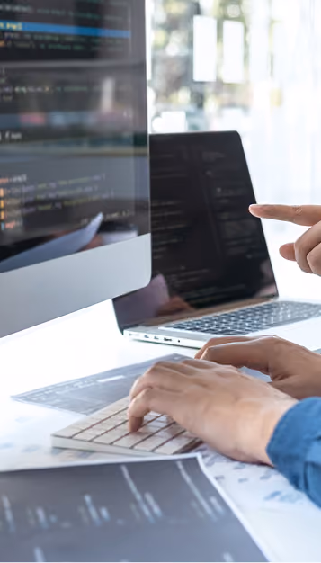 Hands typing on a keyboard with a person pointing at a computer screen showing code.