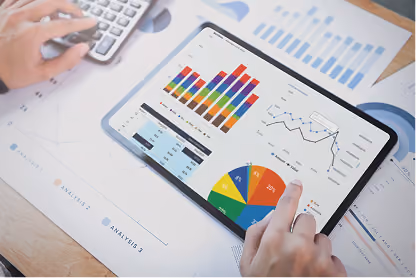 Hands interact with a tablet displaying colorful charts and graphs on a wooden desk with paper charts.