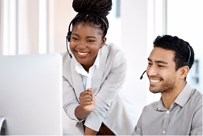 Two smiling customer service representatives wearing headsets looking at a computer screen together.