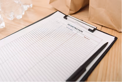 Blank registration form on clipboard with pen on wooden table beside bottles and paper bags.