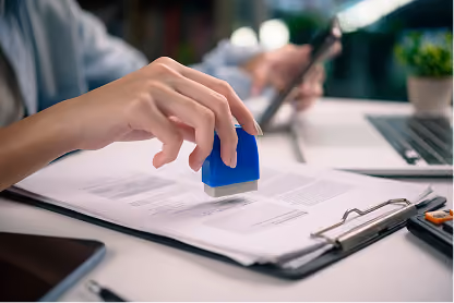 Close-up of a person stamping documents with a blue stamp on a desk with a laptop and clipboard.