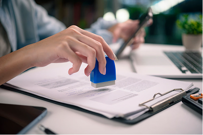 Close-up of a person stamping documents with a blue stamp on a desk with a laptop and clipboard.