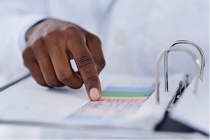 A person in white pointing at a colorful chart or table on paper in a binder.