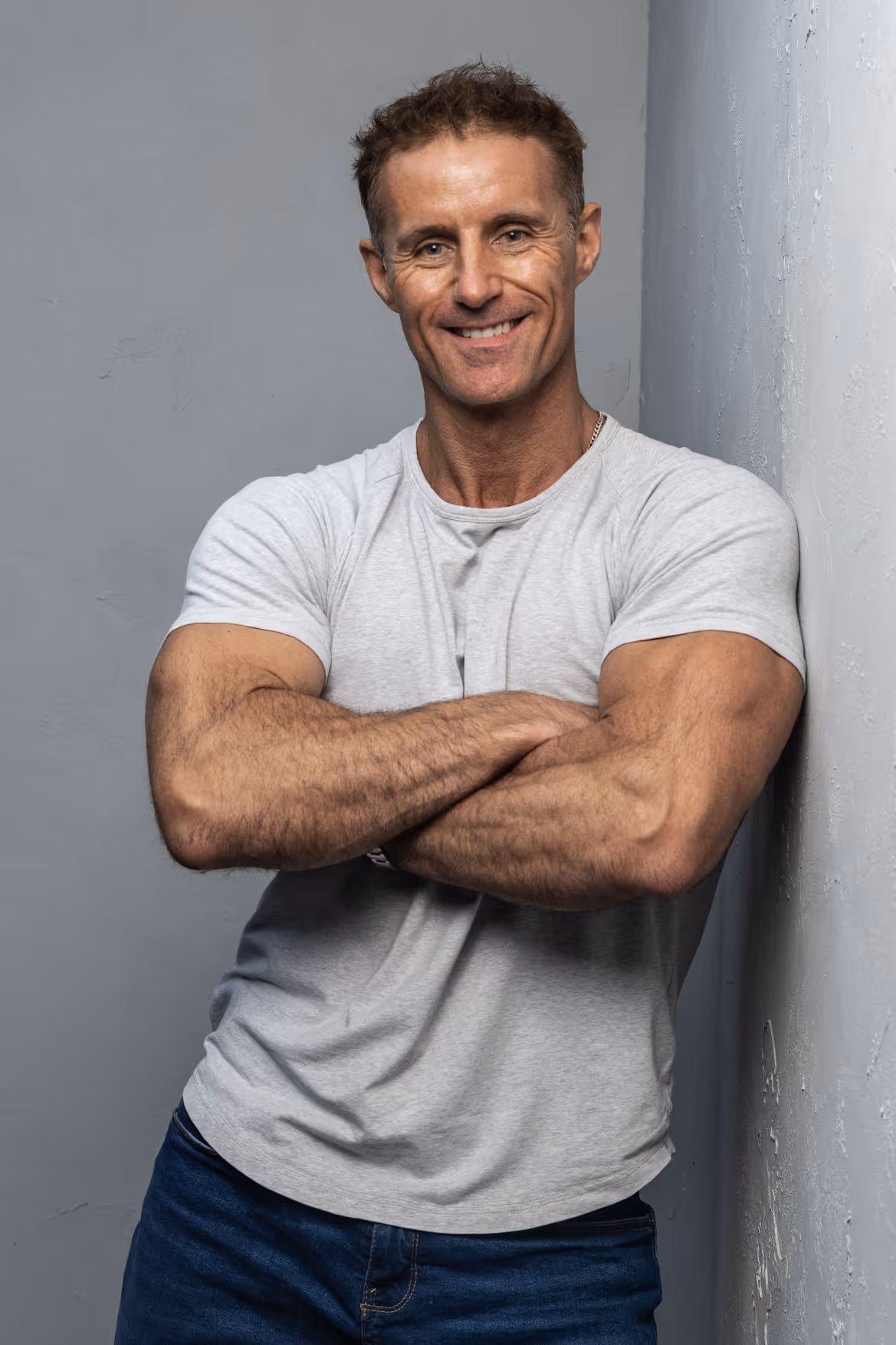Smiling muscular man with crossed arms wearing a gray t-shirt and blue jeans leaning against a wall.