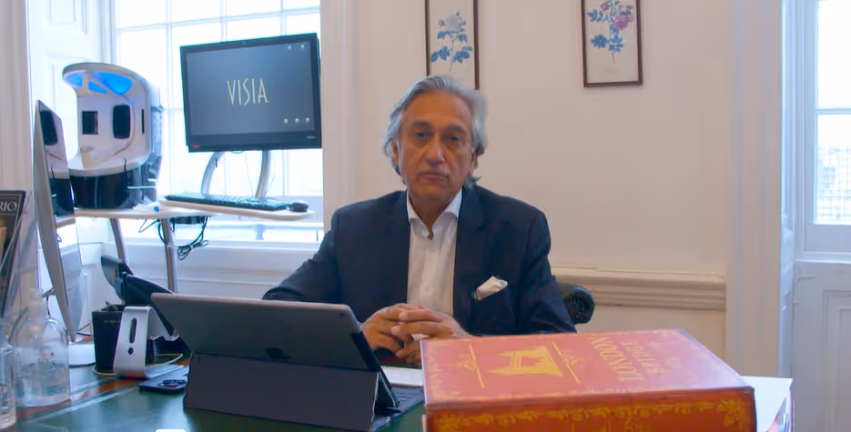 Man in a suit seated at a desk with a tablet, a large red book, and a computer screen showing the word 'VISIA' in the background.