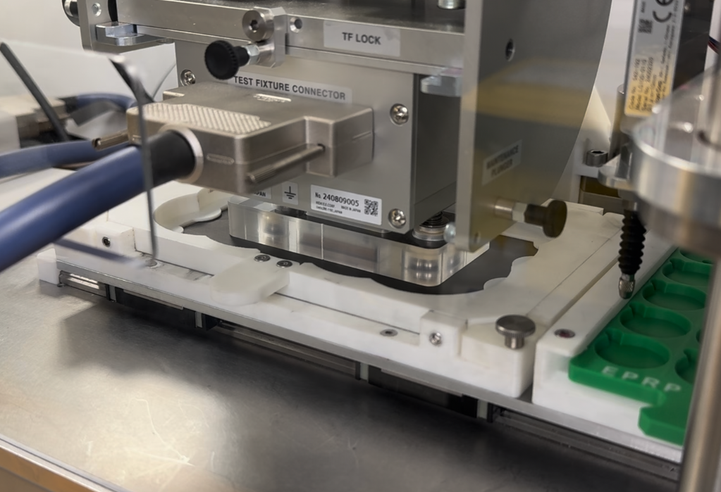 Close-up of a metal and plastic industrial testing device with connectors and labelled parts on a stainless steel surface.