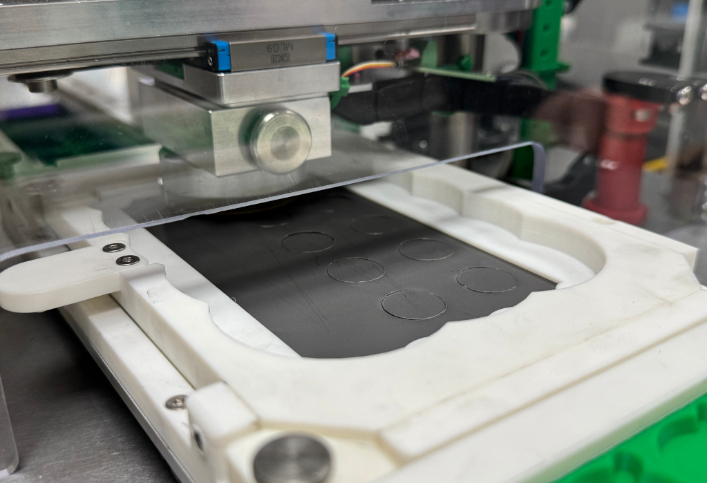 Close-up of an electrode preparation machine cutting circular shapes into a black sheet inside a white and green frame.