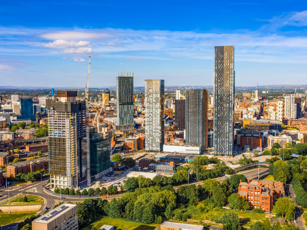 Manchester city skyline featuring modern high-rise buildings, green trees, and a clear blue sky.