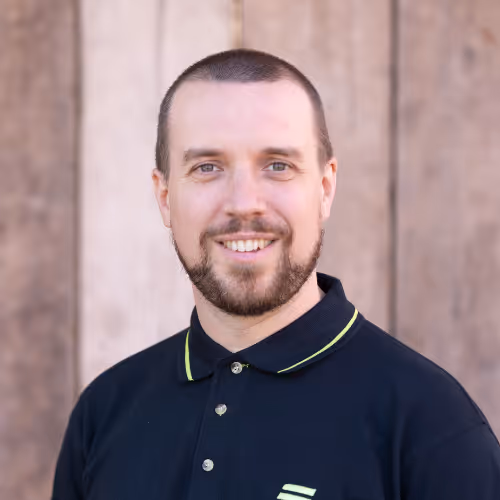 Smiling man with short hair and beard wearing a black polo shirt with a yellow-trimmed collar, standing in front of a wooden background.