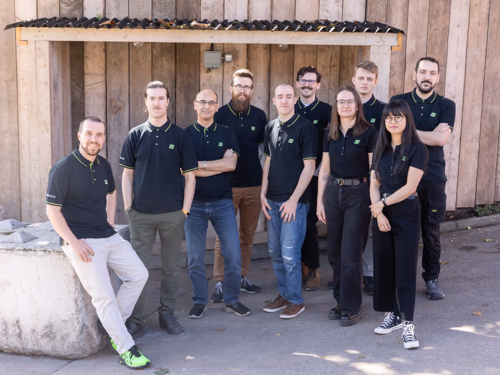 Group of ten people wearing matching black polo shirts with green logos, standing and smiling outdoors in front of a wooden wall.