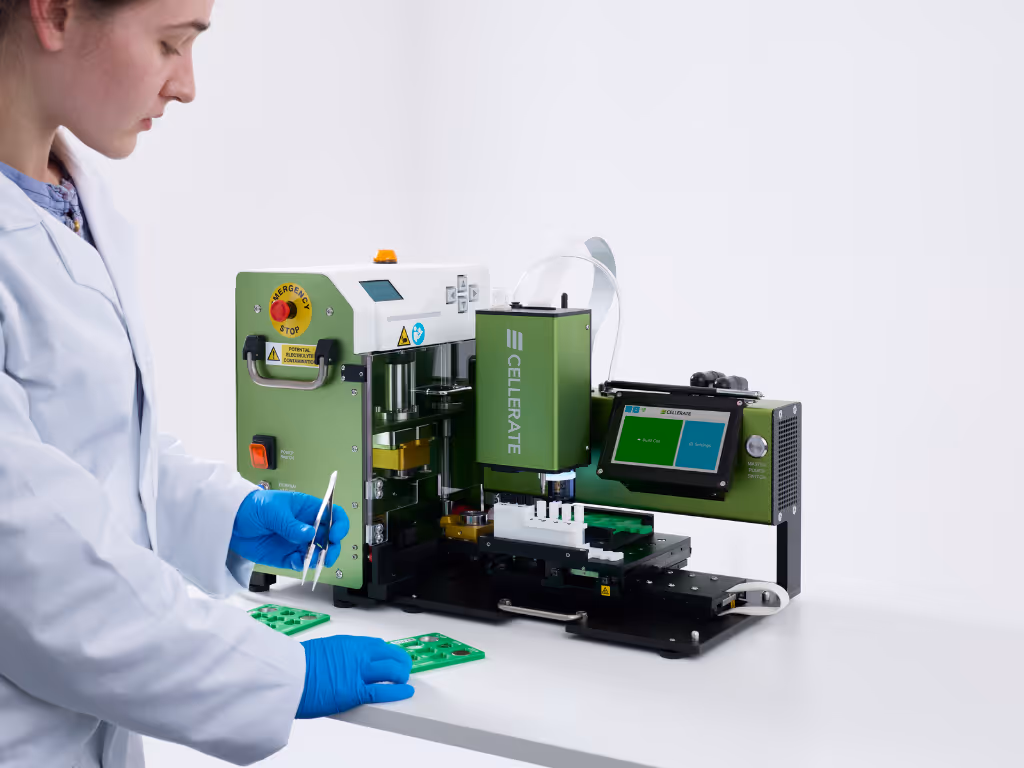 Technician in white coat and blue gloves placing green circuit boards into a green CELLERATE machine with a touchscreen interface on a white table.