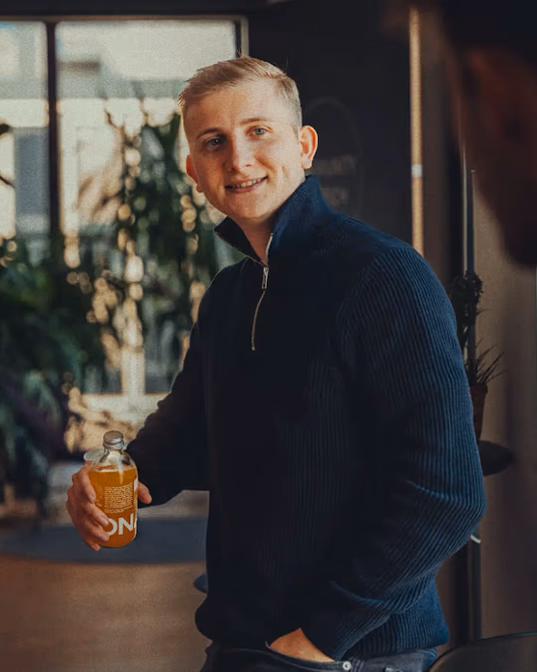 Young man in a dark blue zip-up sweater holding a bottle with an orange drink, smiling indoors with plants in the background.
