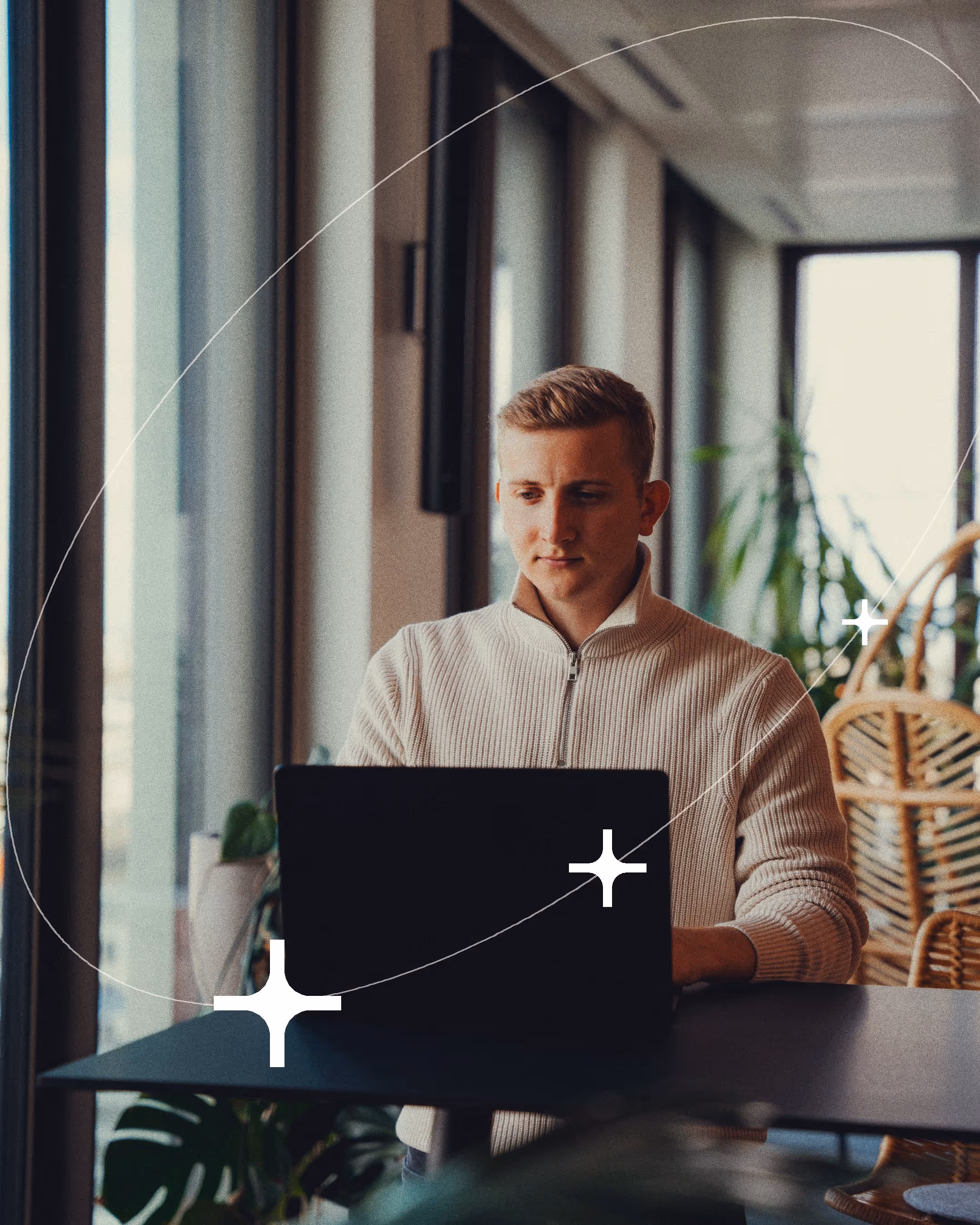 Young man focused on working on a laptop in a bright, modern office with large windows and plants.