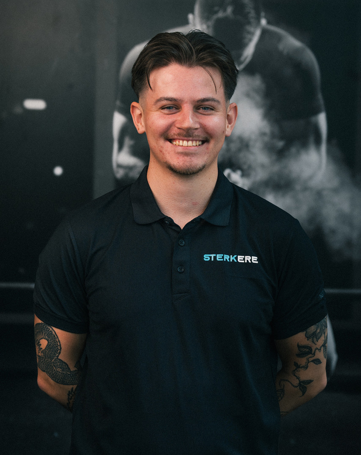 Woman using an elliptical trainer while smiling at a male personal trainer wearing a shirt that reads 'STERKERE Personlig trener'.