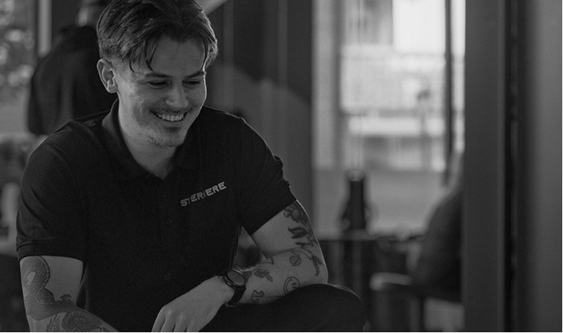 Smiling young man with tattoos on his arms wearing a black polo shirt sitting indoors.