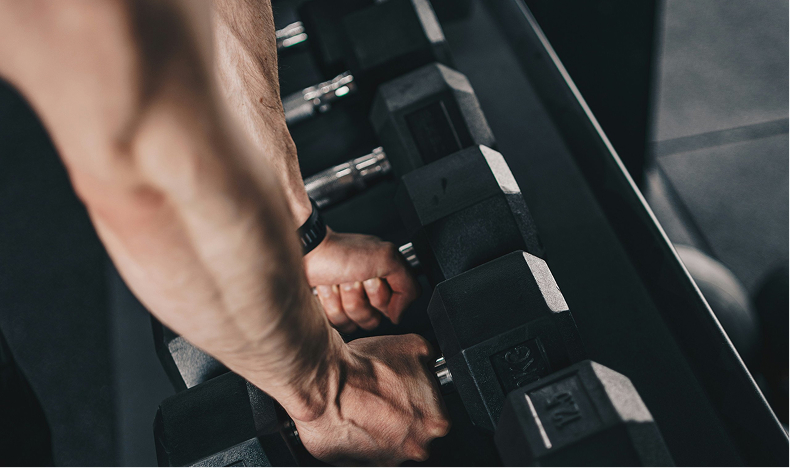 Close-up of muscular arms gripping a dumbbell on a rack in a gym.