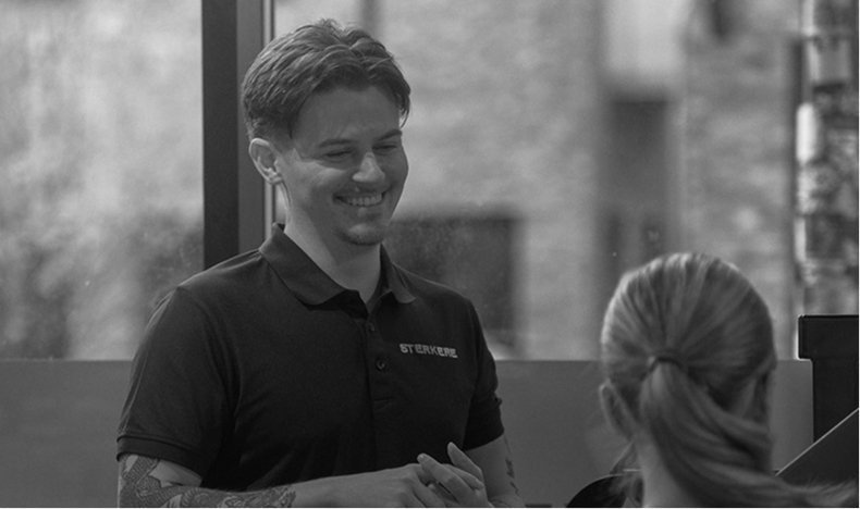 Smiling man in a Sterkere polo shirt talking to a woman with a ponytail in an indoor setting.