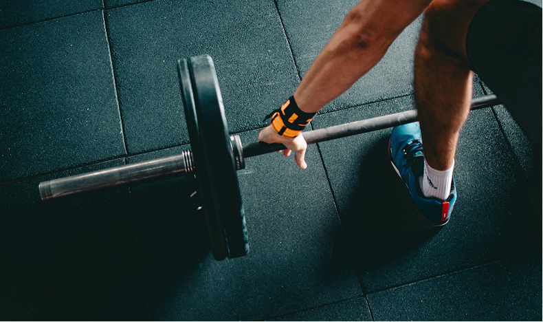 Person wearing wrist wraps gripping a loaded barbell on a gym floor with rubber tiles, preparing to lift.