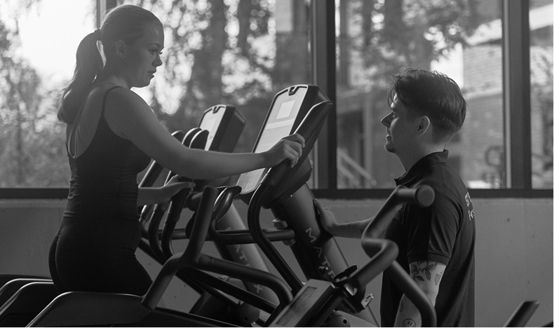 Woman using elliptical machine while a man stands nearby in a gym with large windows.