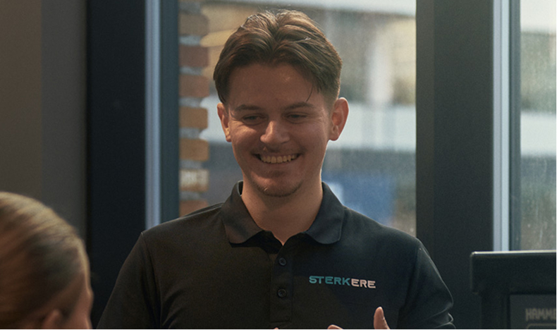 Male personal trainer with tattoos assisting a woman in workout on gym equipment.