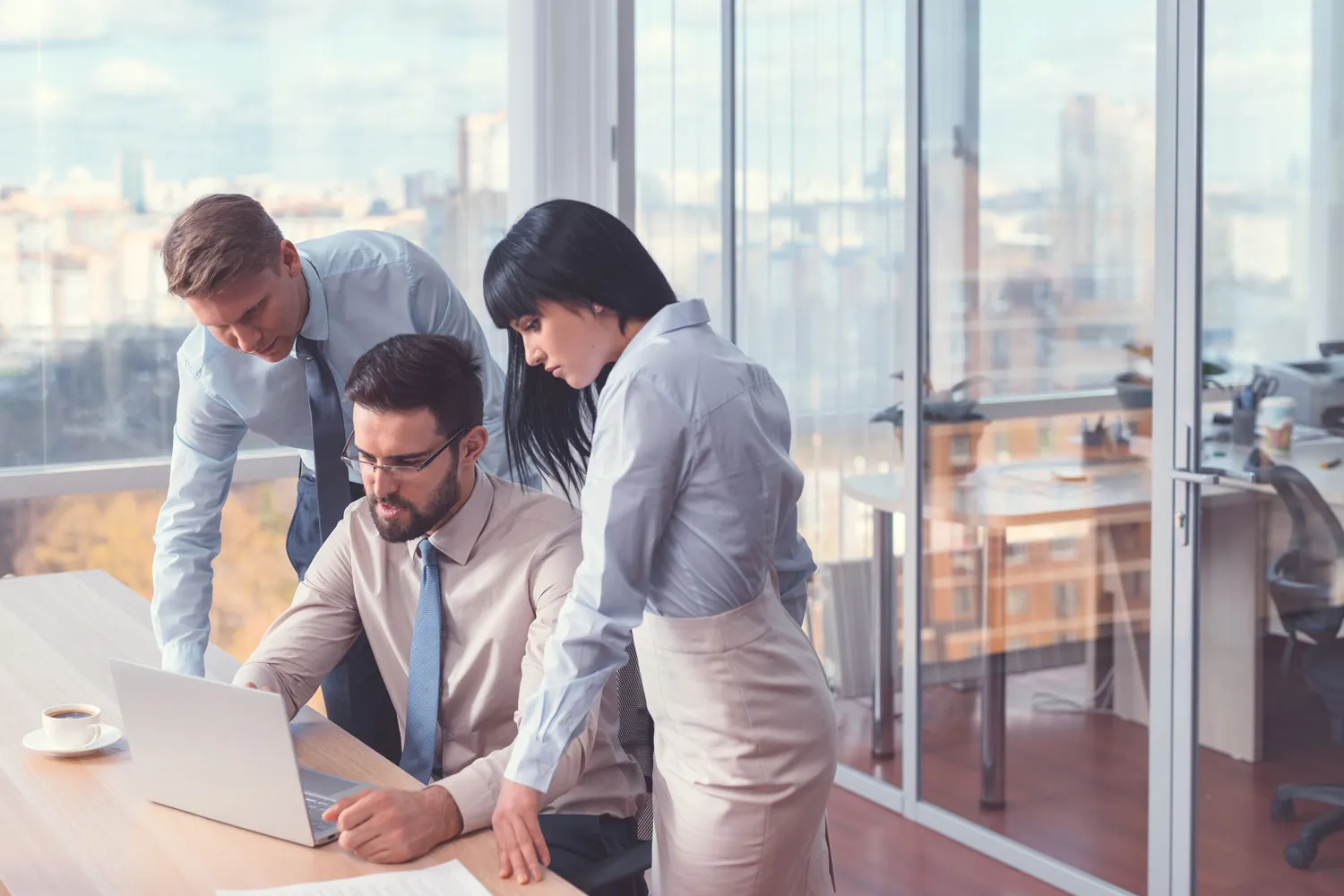 Three business colleagues collaborating over a laptop in a modern office with large windows.