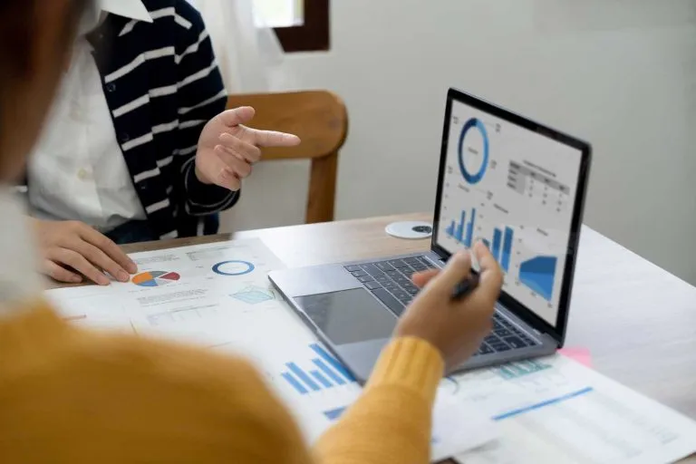 Two people collaborating at a table with financial charts, graphs, and a laptop displaying data visualizations.