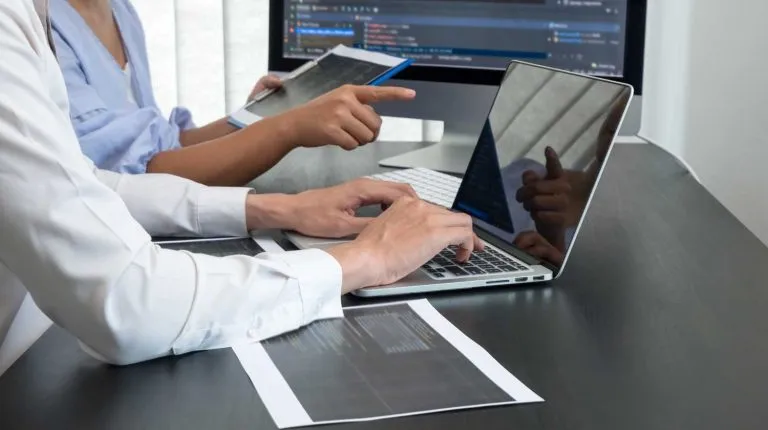 Two people collaborating at a desk with one typing on a laptop and the other pointing at a tablet, with printed documents and a desktop monitor displaying code in the background.