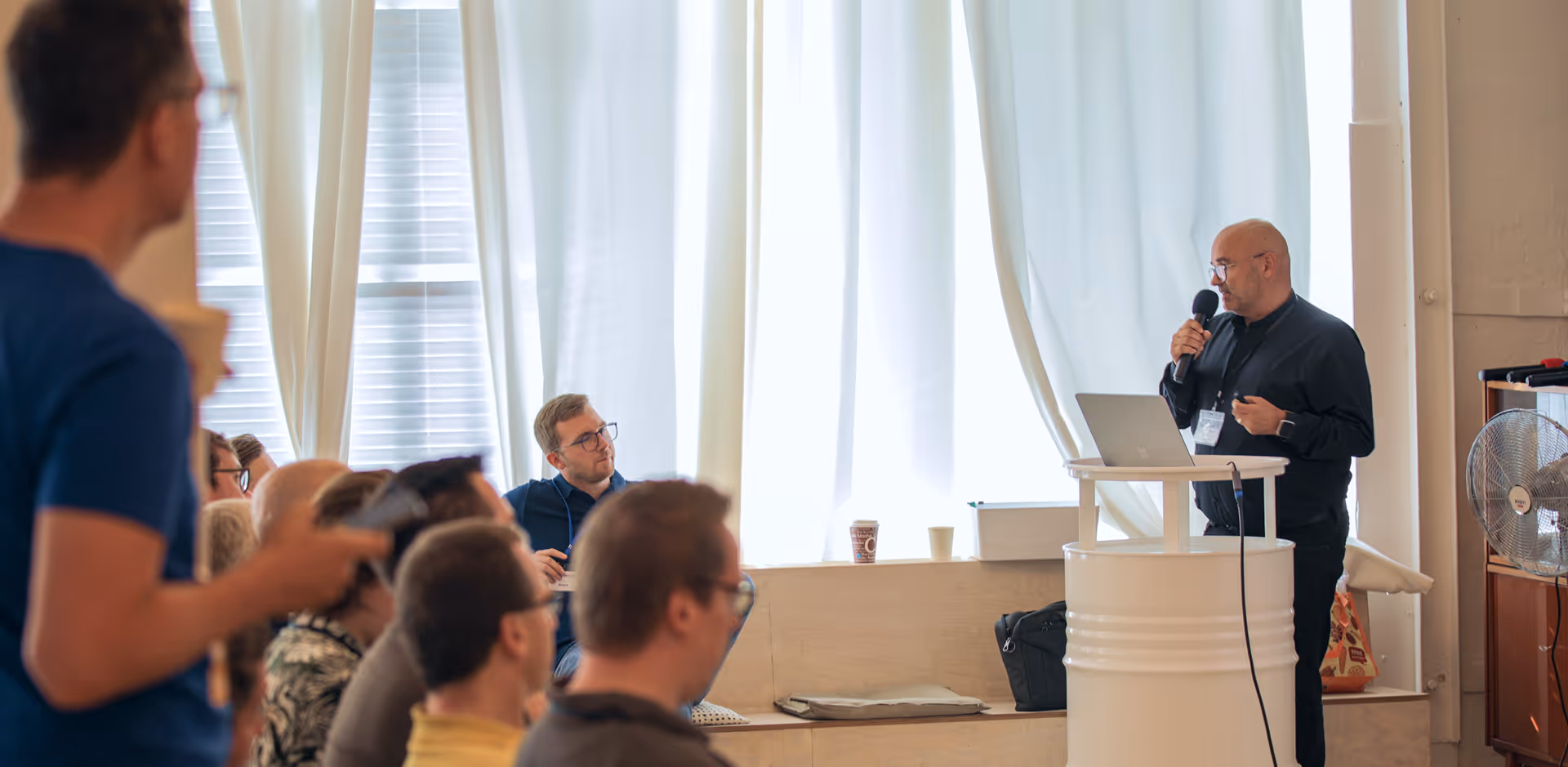 A man giving a speech with a tablet in front of him and holding a microphone while the audience listens. There is a van behind him and the curtains are white
